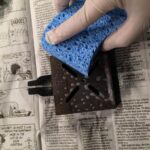 Gloved hand cleaning metal piece with sponge.