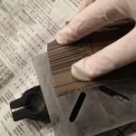 Gloved hand sanding metal piece on newspaper.